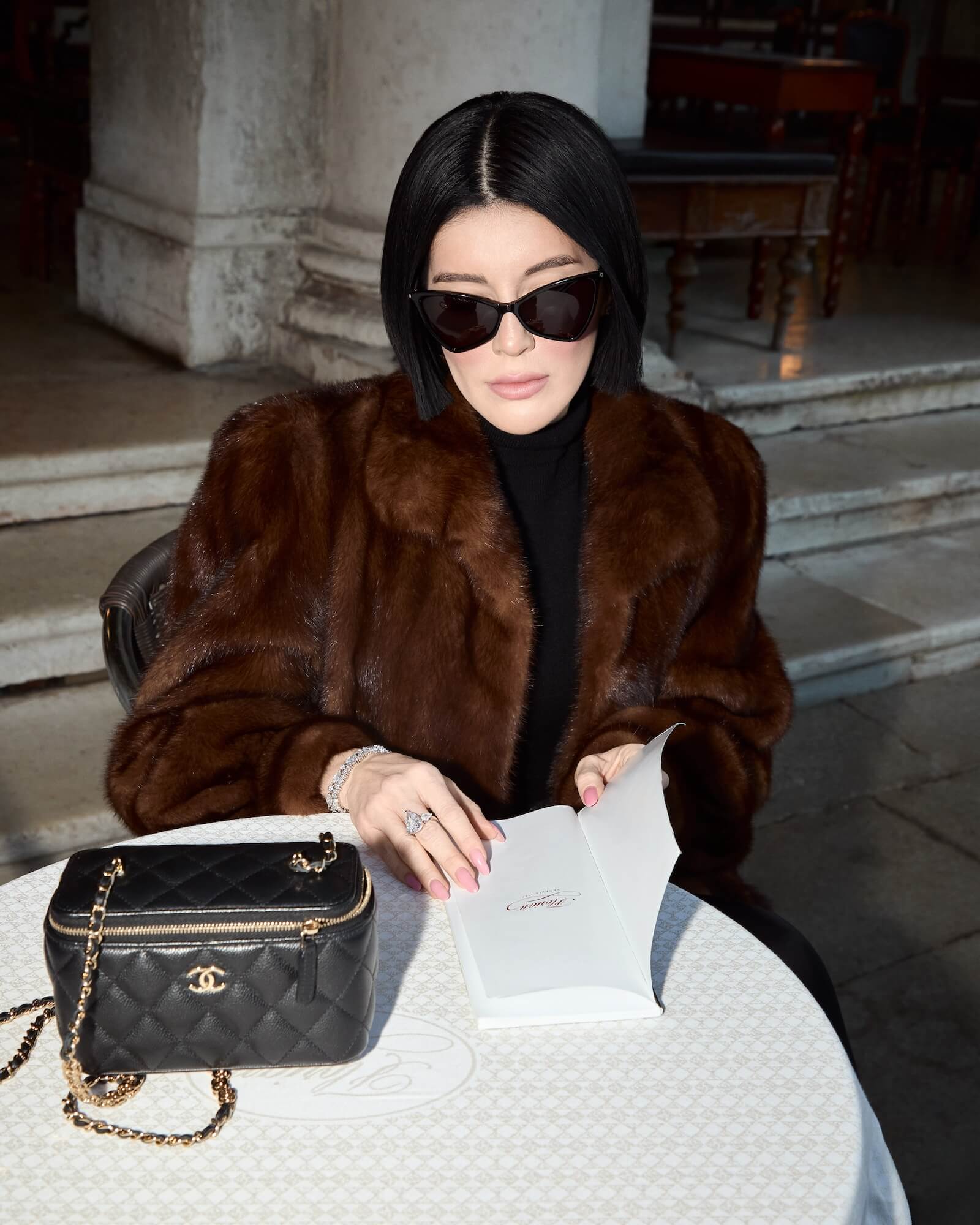 Viv Halil in a fur coat with a Chanel handbag sitting at an outdoor table wearing the Georgina pear cut 3-stone ring