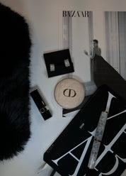Luxury items including The Amira pavé hoop earrings, a lip gloss, a Dior round box, a black clutch, and a fountain pen, on a white surface with a Harper's Bazaar in the background.
