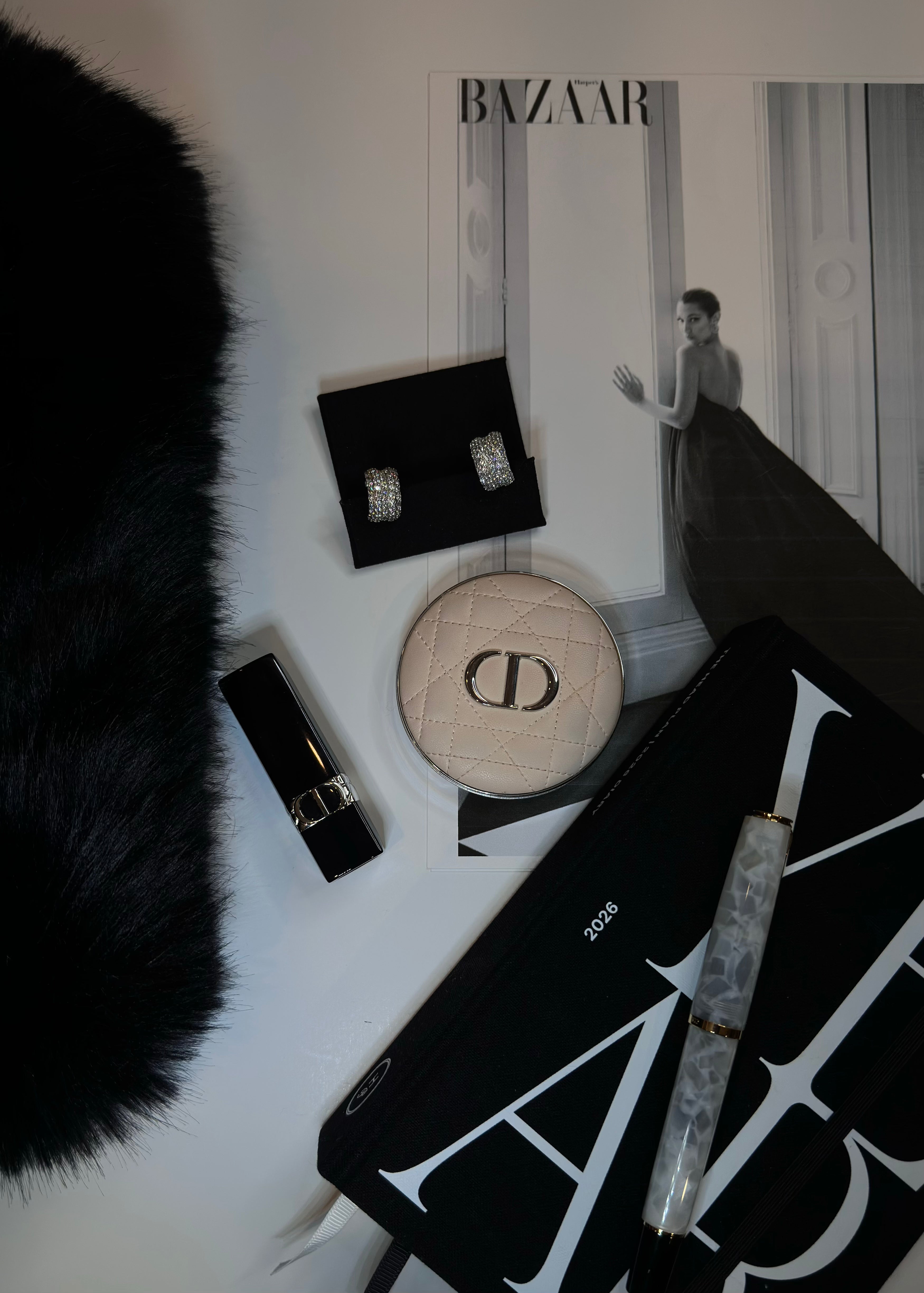Luxury items including The Amira pavé hoop earrings, a lip gloss, a Dior round box, a black clutch, and a fountain pen, on a white surface with a Harper's Bazaar in the background.