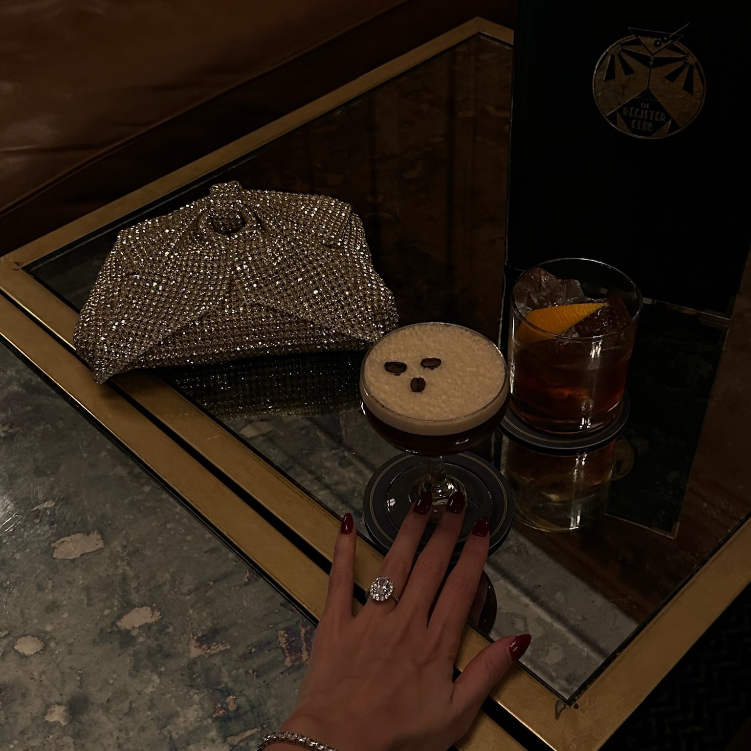 Model's hand with a statement ring on a table with a handbag and drinks, 3mm petite luxxe tenné tennis bracelet on the wrist.