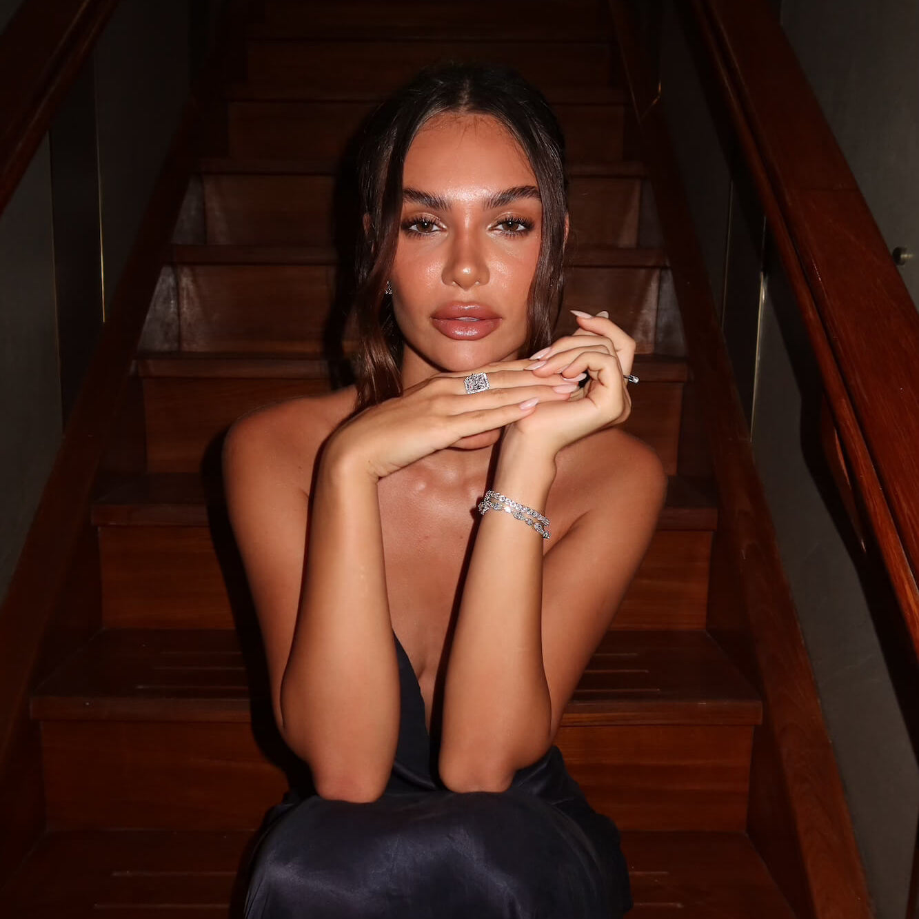 Model in a dark  dress sitting on wooden stairs wearing the Aurora Sterling silver emerald cut halo ring and a set of VH bracelets