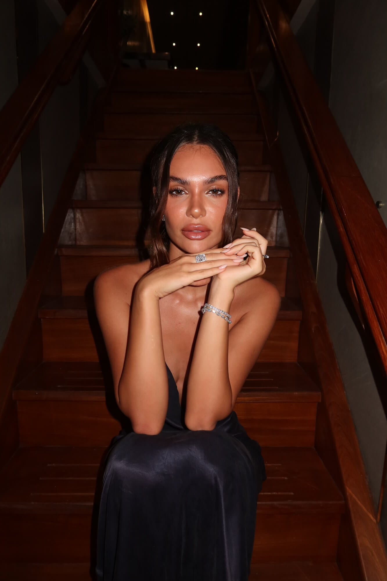 Model in a dark  dress sitting on wooden stairs wearing the Aurora Sterling silver emerald cut halo ring and a set of VH bracelets
