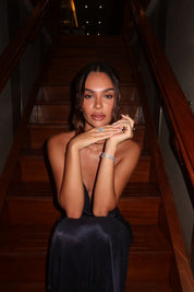 Model in a dark  dress sitting on wooden stairs wearing the Aurora Sterling silver emerald cut halo ring and a set of VH bracelets