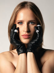 Woman wearing black leather gloves with diamond alternative rings on a neutral background.