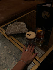 Hand with The Mon Cheri Ring by Viviana Halil on a table with a handbag and drinks.