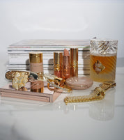 Cosmetic items, including lipsticks, a compact mirror, and a perfume bottle on a reflective surface, along with VH essential jewelry pieces.