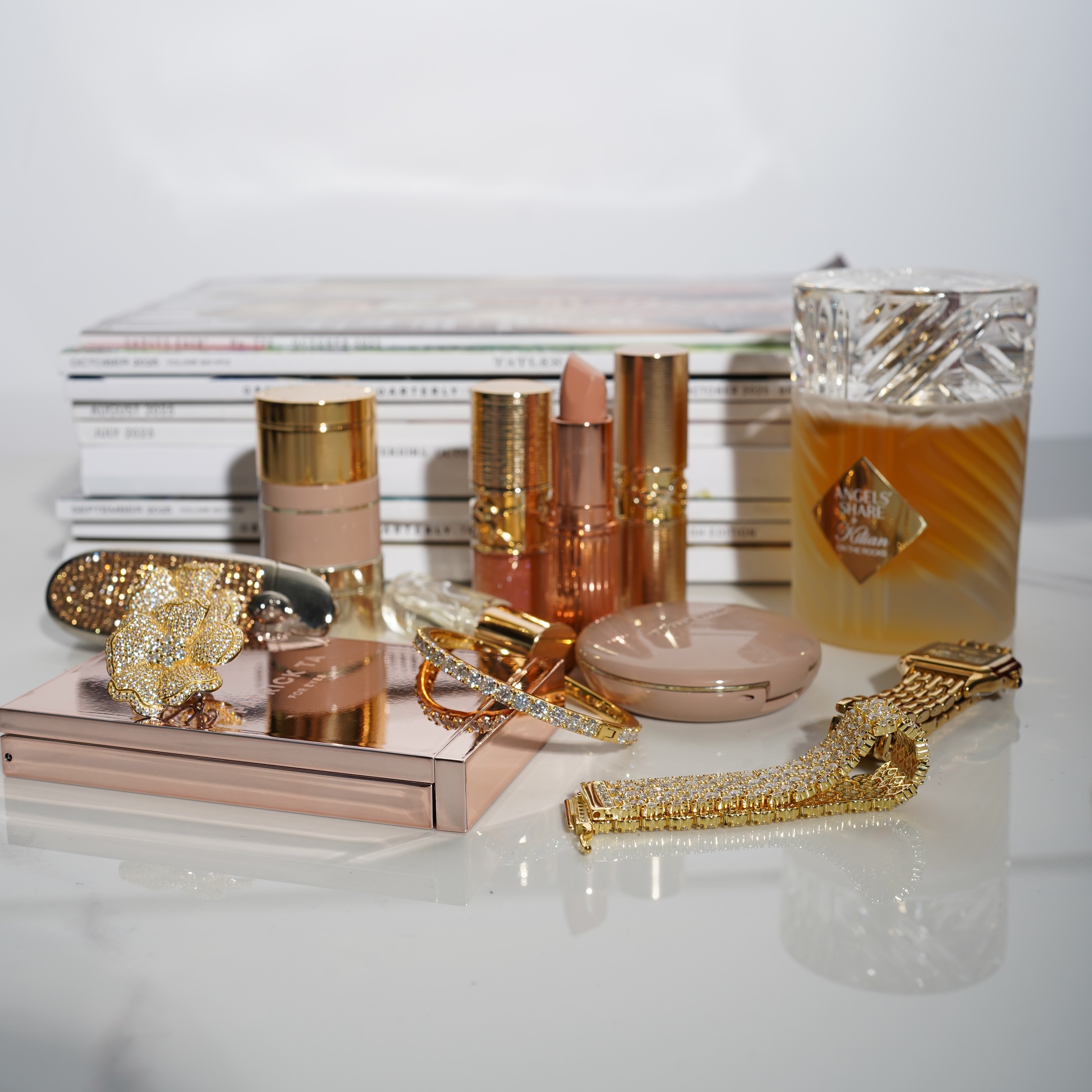 Cosmetic items, including lipsticks, a compact mirror, and a perfume bottle on a reflective surface, along with VH essential jewelry pieces.