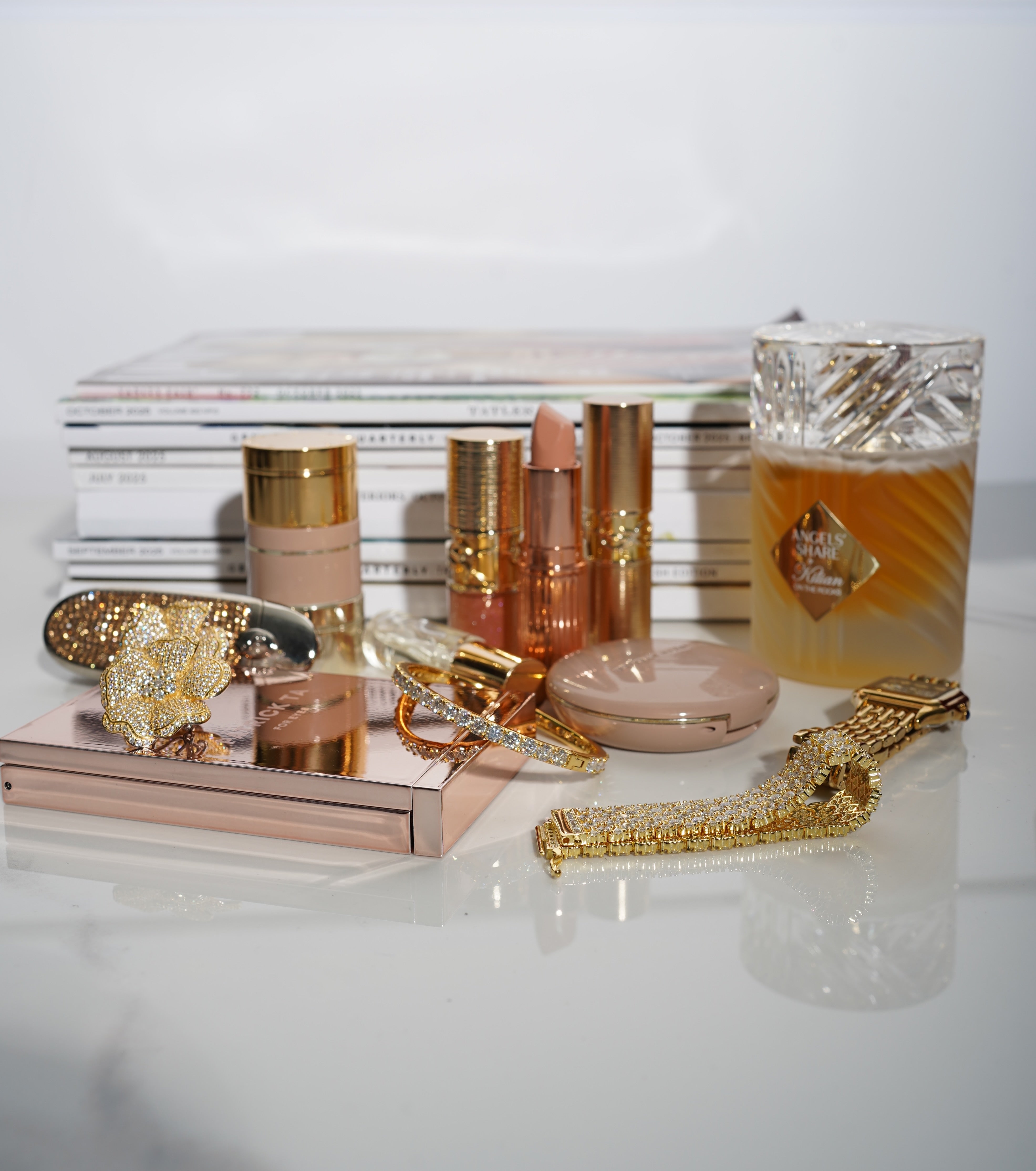 Cosmetic items, including lipsticks, a compact mirror, and a perfume bottle on a reflective surface, along with VH essential jewelry pieces.